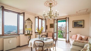 A dining area with a round table, chairs, a chandelier, and a view through an open window. - Lenno Il Guscio (Tremezzina)
