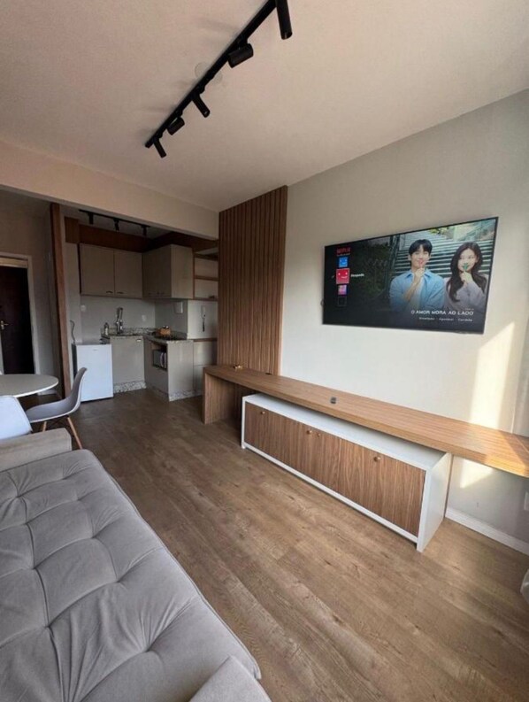 Une chambre moderne avec un lit, un bureau, un téléviseur fixé au mur et un coin cuisine à l’arrière-plan.