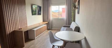 Ein modernes Wohnzimmer mit einem runden Esstisch, zwei weißen Stühlen, einer Couch, einer hölzernen Bank und einem großen Fenster mit Vorhängen.
