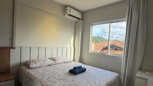 Ein Schlafzimmer mit einem Bett, einer Klimaanlage, einem Fenster mit Vorhängen und Blick auf Gebäude.