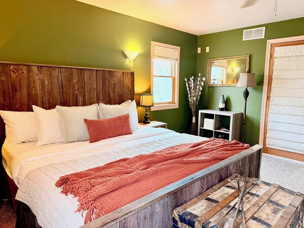 A bedroom with a wooden headboard, a bed with white bedding and a red throw, a nightstand with a lamp, a mirror, and a vase with flowers.