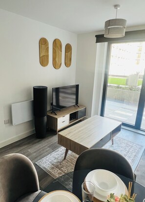 Ein modernes Wohnzimmer mit Flachbildfernseher, Couchtisch aus Holz, Esstisch mit Stühlen und einem großen Fenster mit Blick ins Freie.