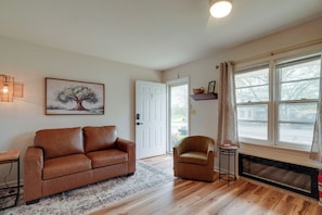 A living room with a brown leather sofa, a brown armchair, a patterned rug, a fireplace, a large window with curtains, and a painting of a tree.
