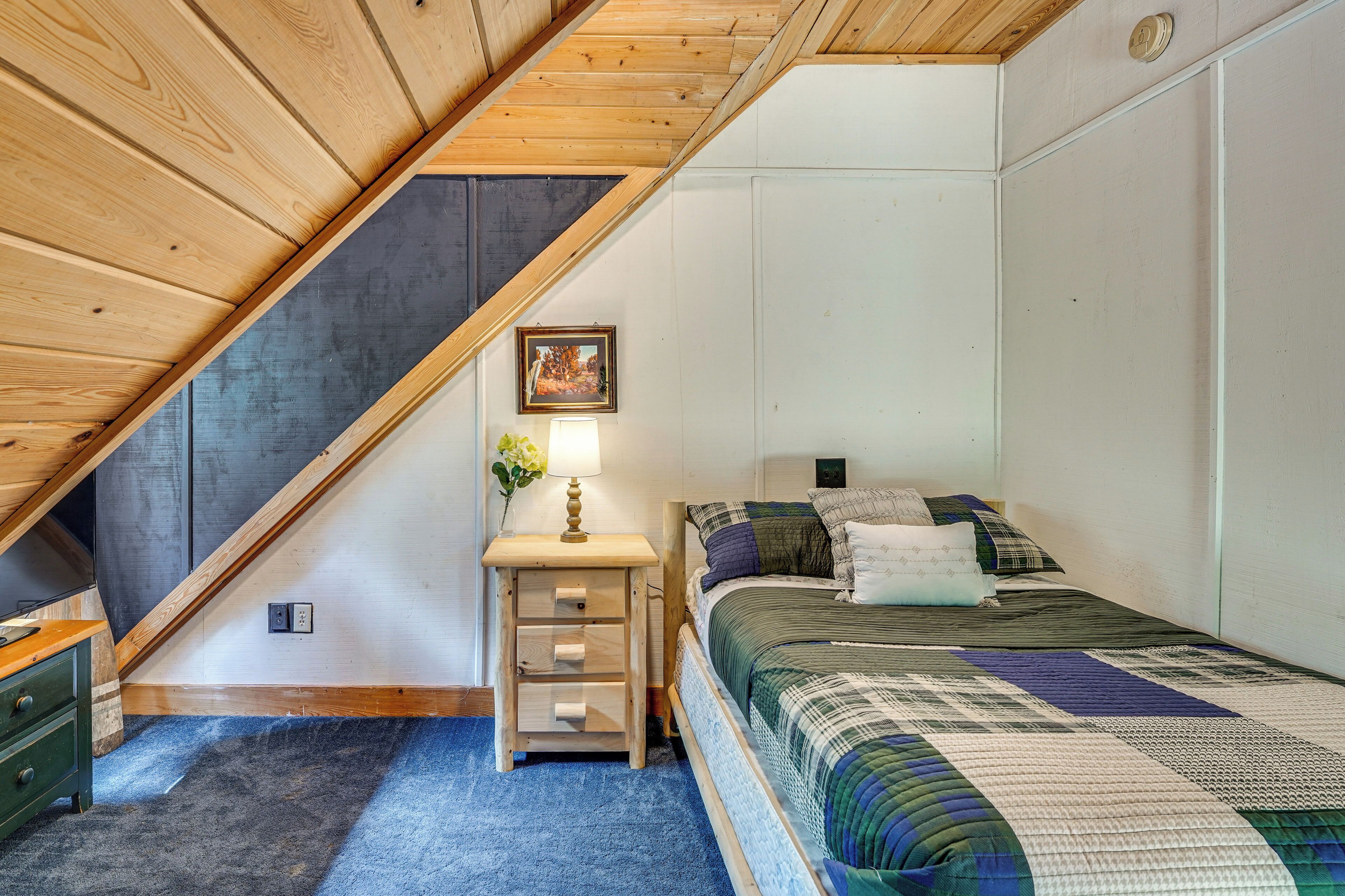 Une chambre avec un plafond en pente, un lit recouvert d’une courtepointe, une table de chevet avec une lampe et un tableau accroché au mur.