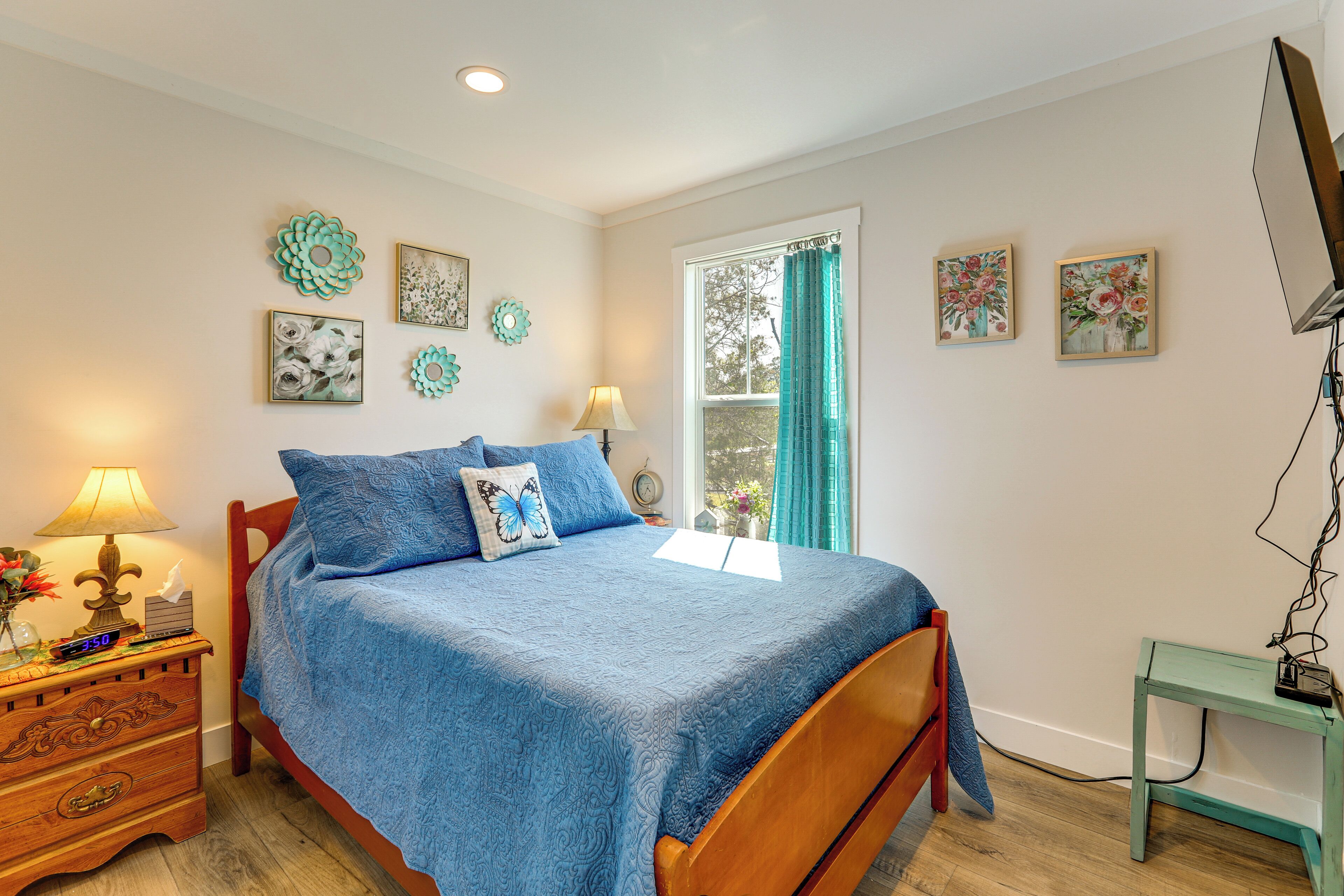 A bedroom with a bed, bedside table, lamp, wall art, and a window with curtains.