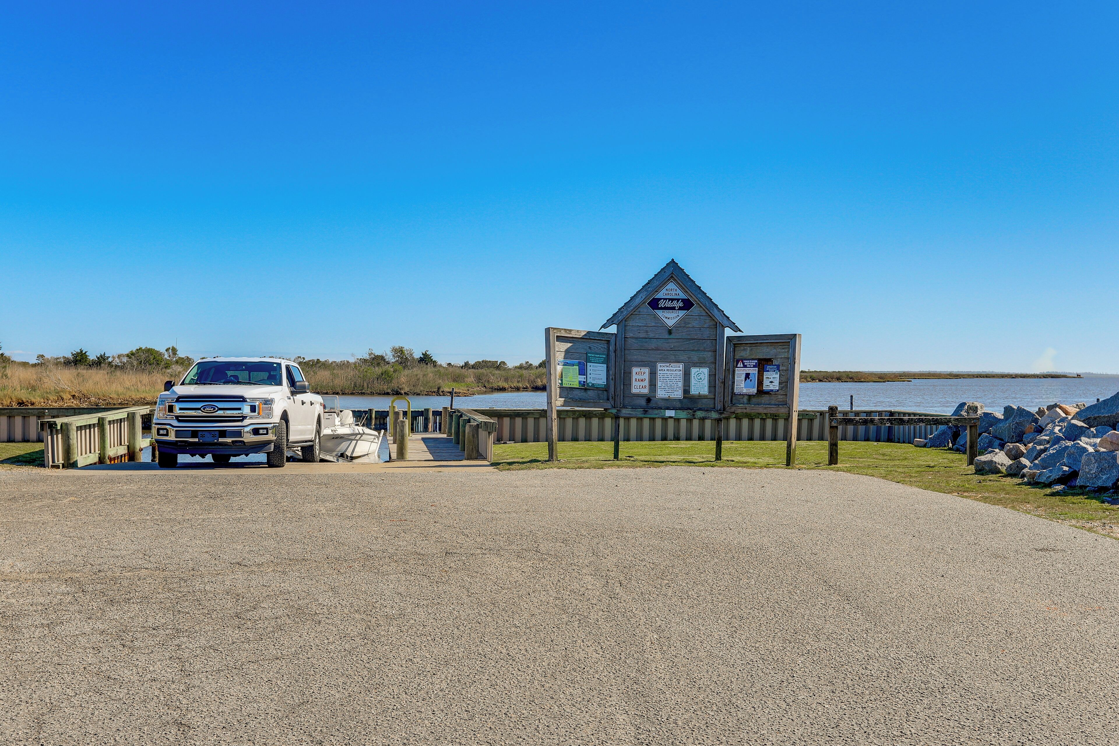 A gravel parking area with a white pickup truck, a wooden structure with a sign, and a body of water in the background.