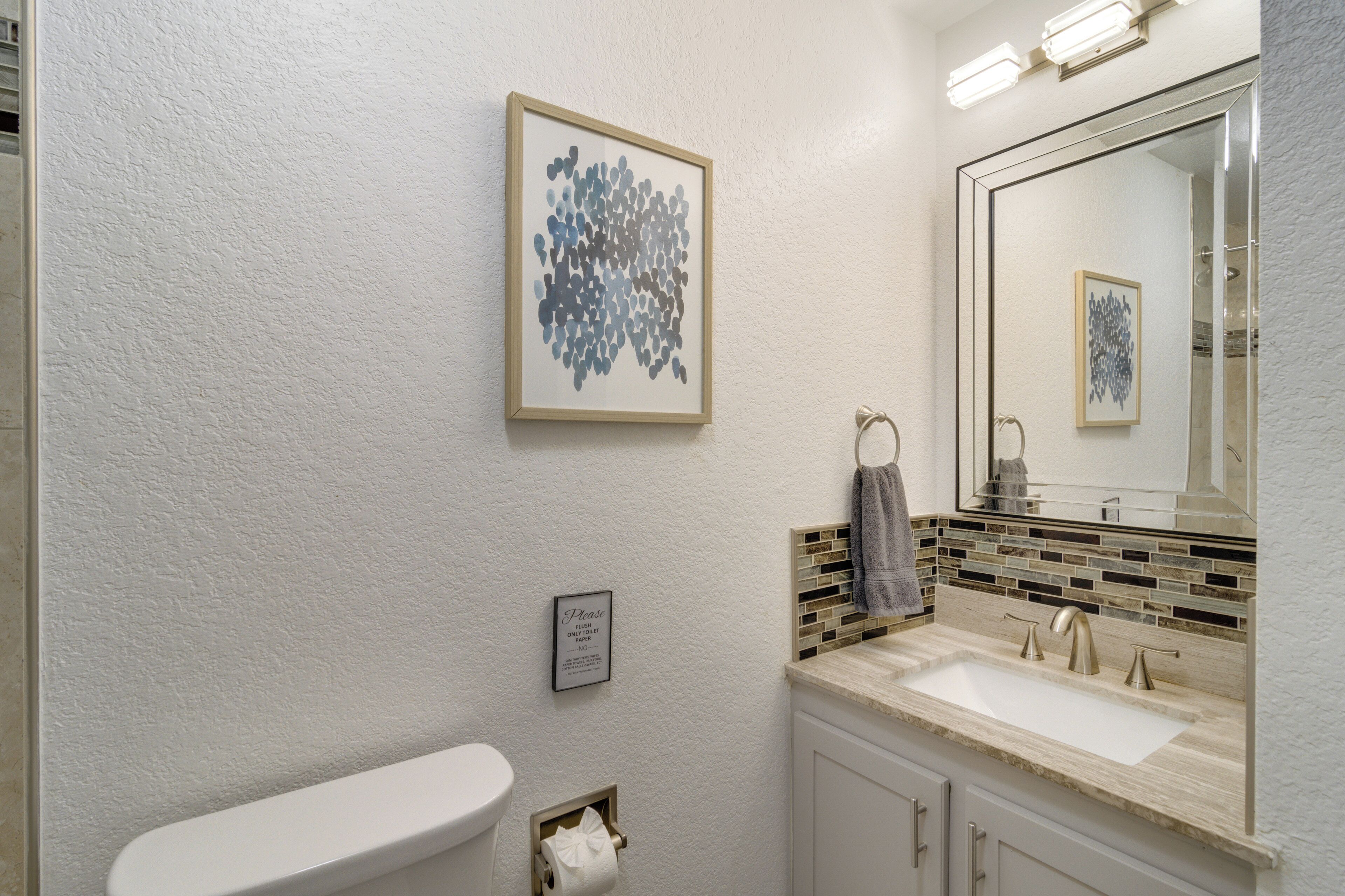 Un baño con inodoro, lavamanos, espejo y una obra de arte abstracto enmarcada en la pared.