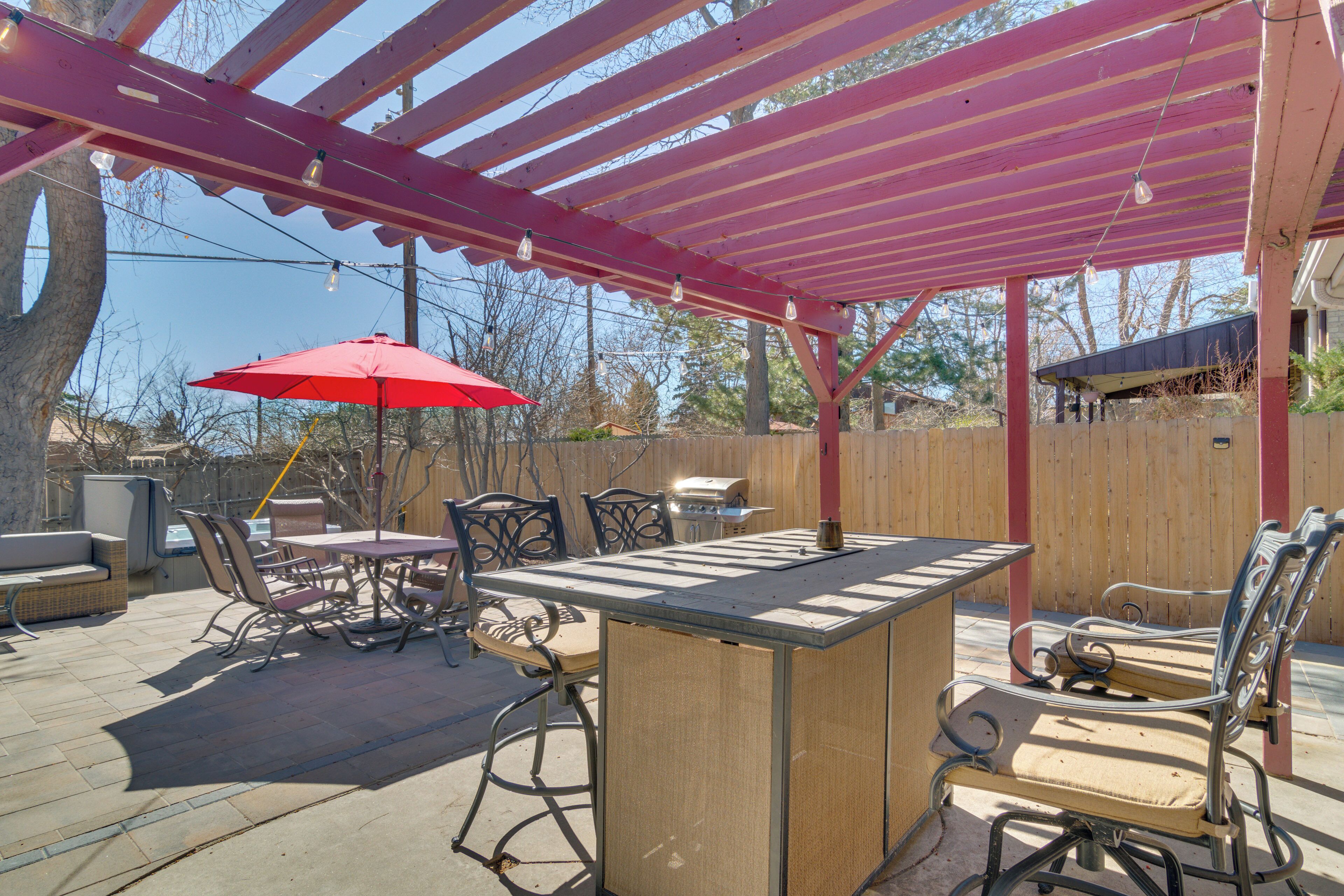 An outdoor patio with a wooden table and chairs, a red umbrella, and a pergola with string lights.