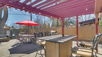 An outdoor patio with a wooden table and chairs, a red umbrella, and a pergola with string lights.