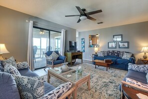 A living room with a ceiling fan, a glass coffee table, a sofa, a desk, and a view through a sliding glass door. - Gwynn's Island Gem Private Beach & Bay Views (Gwynn)