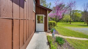 Une maison en bois de couleur brune, avec une porte blanche, un paillasson et un jardin où les arbres sont en fleurs.