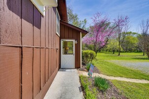 A brown wooden house with a white door, a welcome mat, and a garden with blooming trees.