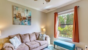 Ein Wohnzimmer mit einem beigefarbenen Sofa, einer blauen Bank, einem Bild an der Wand, einem Fenster mit Vorhängen und einer Lampe.