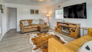 A living room with a brown leather sofa, a wooden cabinet, a large TV mounted on the wall, and a cowhide rug. - Near Ocean & Redwood National Park! Home w/ Yard (Crescent City)