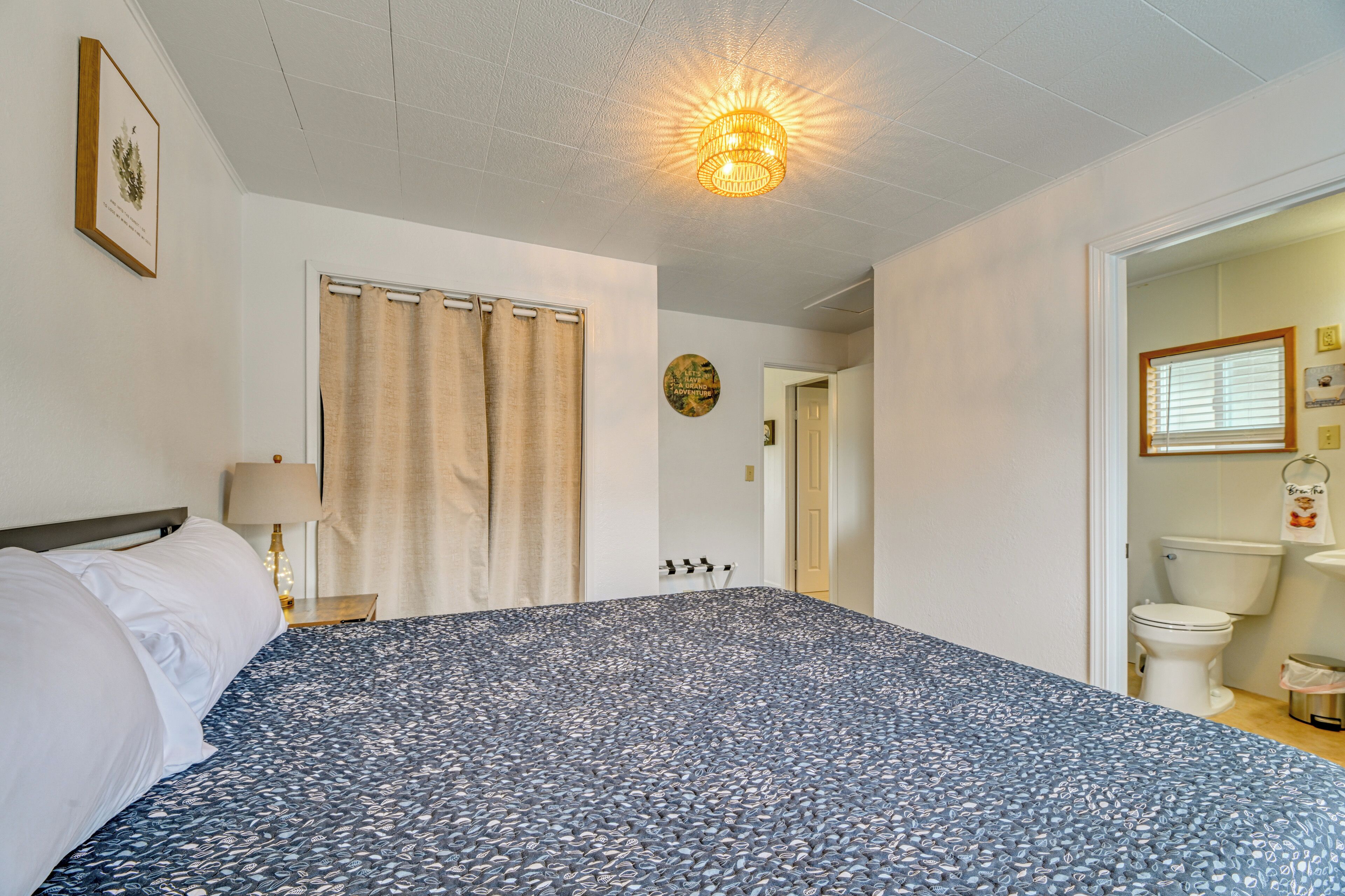 A bedroom with a bed, a nightstand, a lamp, a ceiling light fixture, a door, and a view into the bathroom.