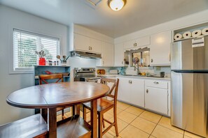Une cuisine avec des meubles blancs, un réfrigérateur en acier inoxydable, une table à manger en bois et une fenêtre avec des stores.