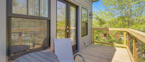 A deck with a table and chair, large windows, and a view of trees and a fence.