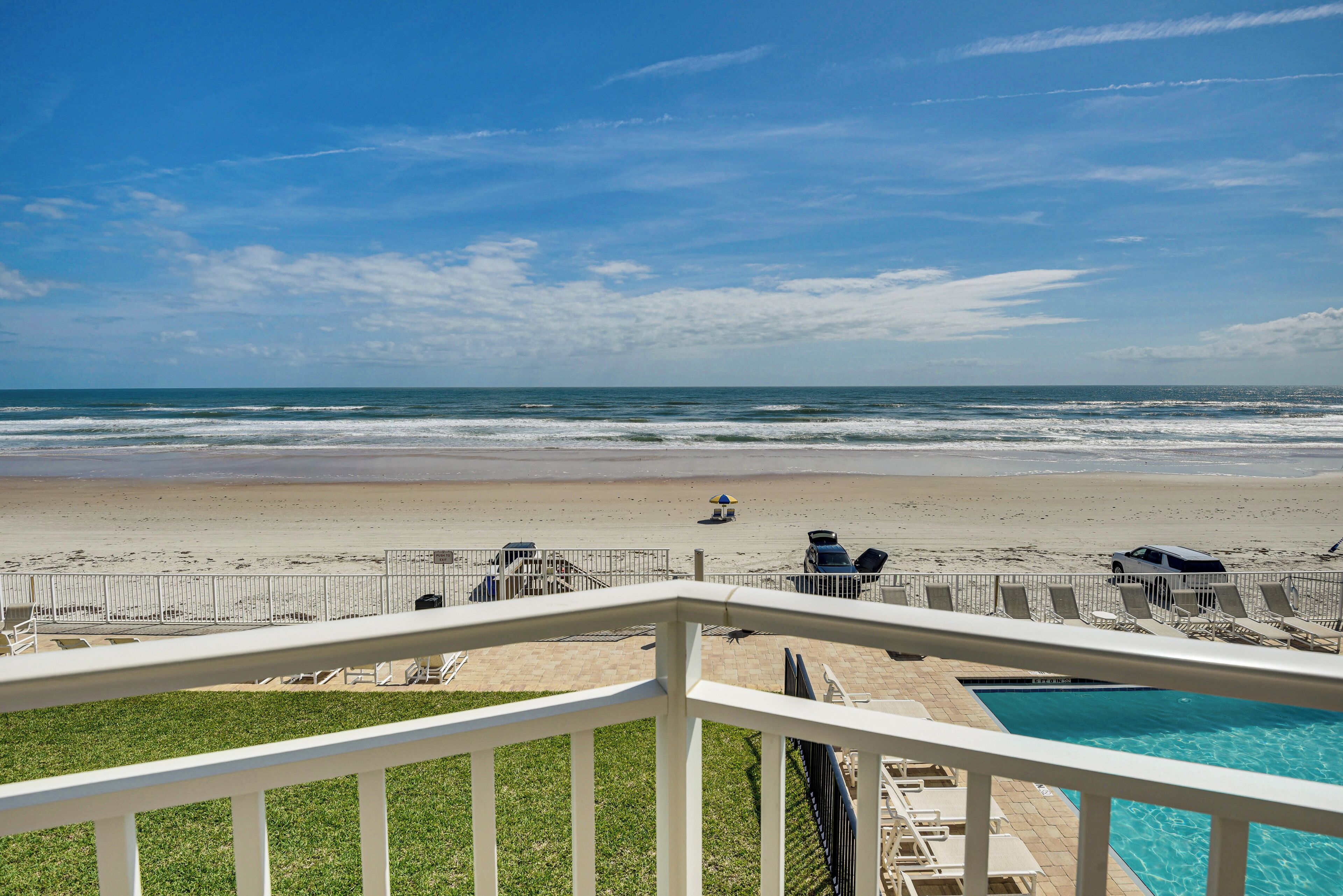 A beach view from a balcony with a pool, beach chairs, and umbrellas.