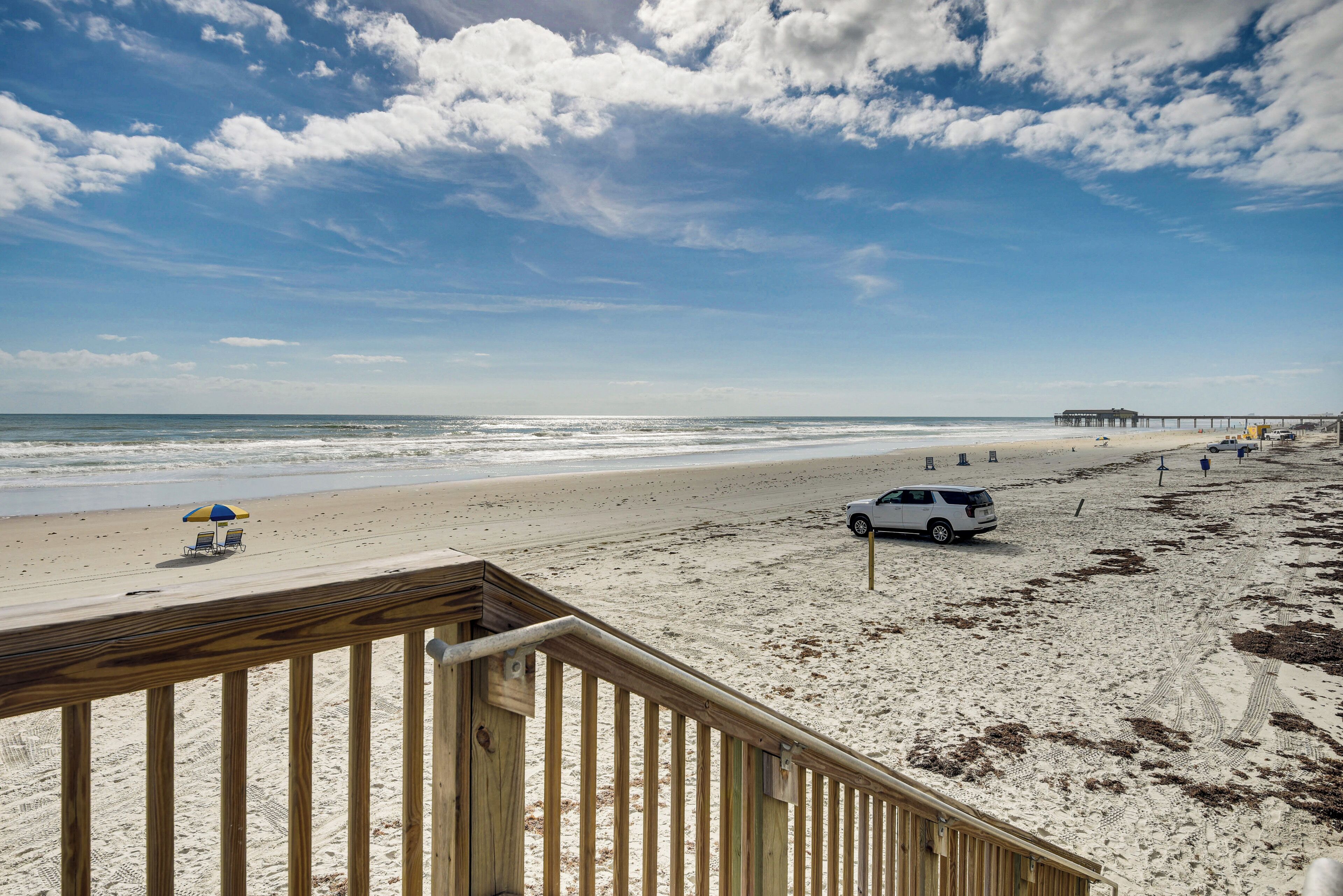 A beach with a wooden deck, a white car, and a pier in the distance.