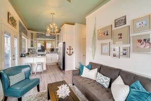 Ein modernes Wohnzimmer mit einem grauen Sofa, einem Türkis-Sessel und einem Couchtisch. Im Hintergrund ist eine Küche mit weißen Schränken und einem Essbereich zu sehen.