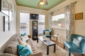 A living room with a sofa, armchairs, a coffee table, and a TV. There is a view of a lake and a dock outside.