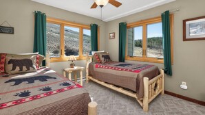 Une chambre avec deux lits, un ventilateur de plafond, une fenêtre donnant sur un paysage montagneux et une petite table avec une lampe.