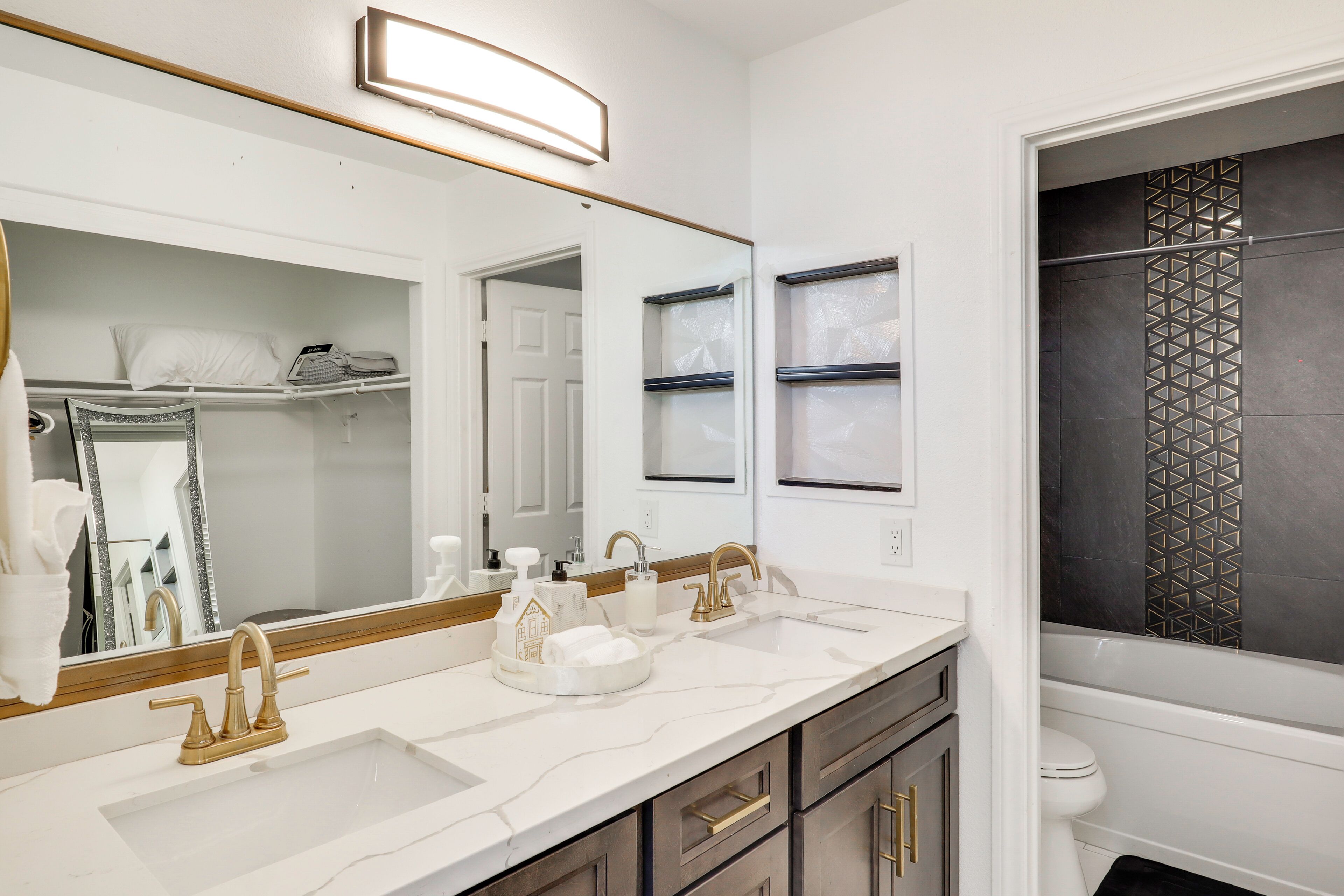 A bathroom with a white vanity, a large mirror, a gold faucet, and a shower with a geometric tile design.