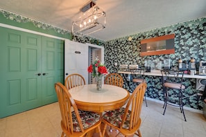 Een eetruimte met een ronde tafel, houten stoelen, een vaas met bloemen, een keukenblad met apparatuur en een groene deur.