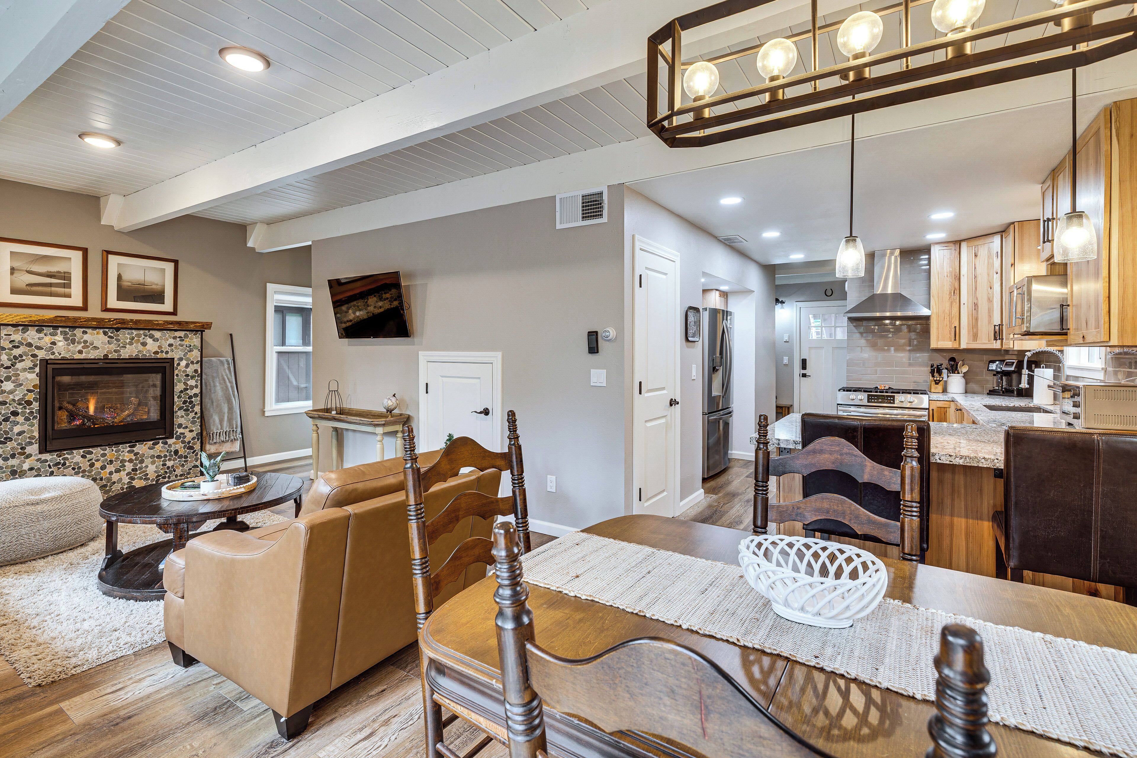 A spacious living area with a fireplace, a dining table with chairs, a kitchen with wooden cabinets, and a ceiling with recessed lighting.
