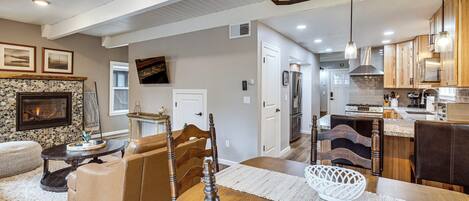A spacious living area with a fireplace, a dining table with chairs, a kitchen with wooden cabinets, and a ceiling with recessed lighting.
