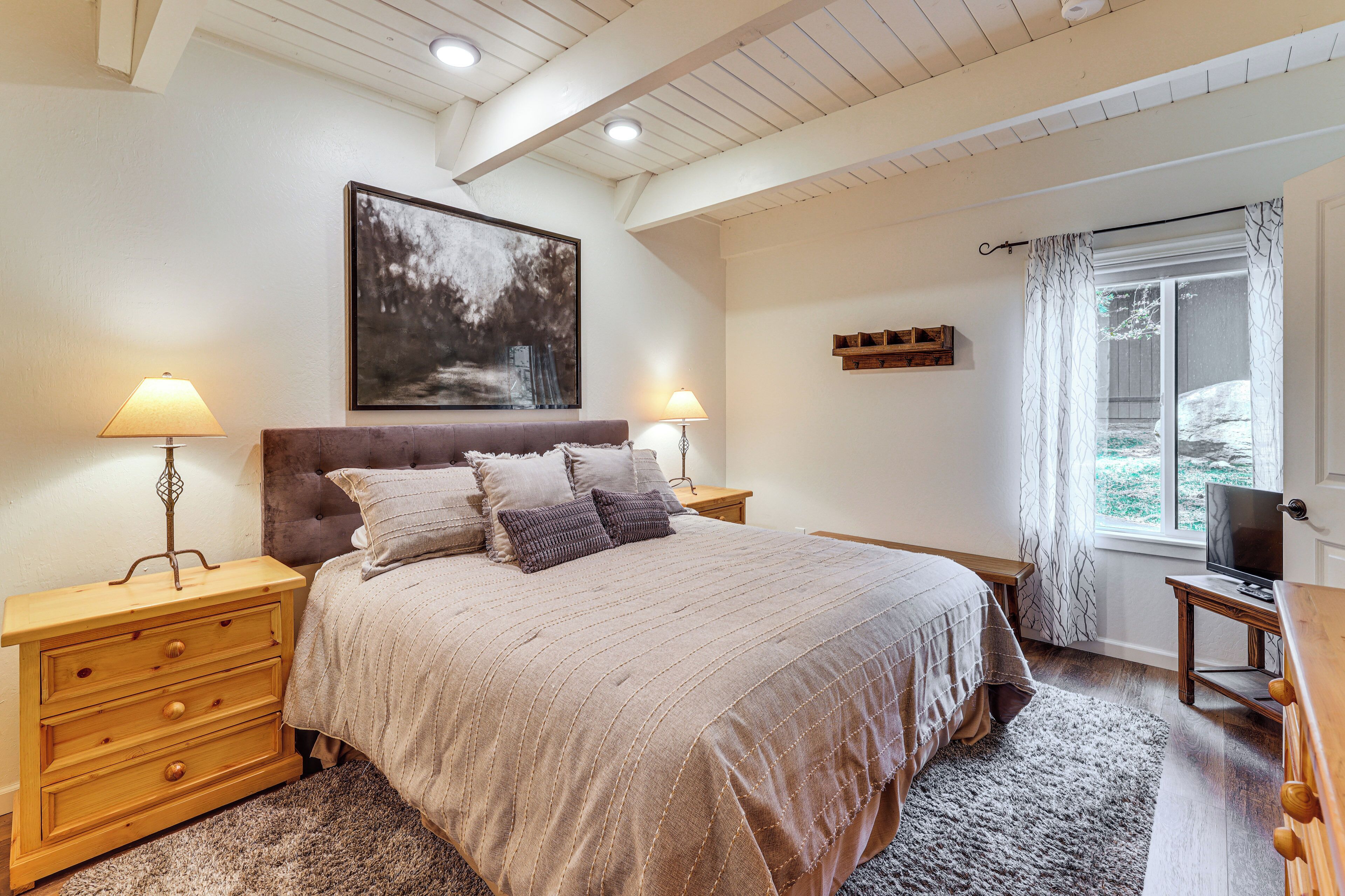 A bedroom with a bed, two bedside tables with lamps, a large painting on the wall, a window with curtains, and a television on a stand.