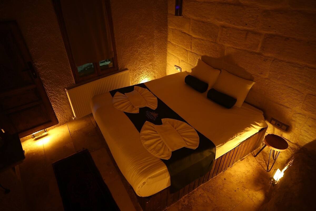 Una cama con almohadas y una colcha, un radiador, una mesita y una pared de piedra.