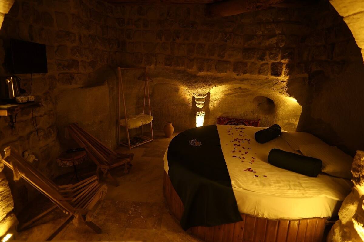 Una habitación con paredes de piedra, una cama, una silla colgante y una mesita con un florero.