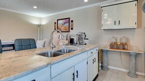 Una cocina con encimera de granito, pileta de acero inoxidable, armarios blancos y un estante decorativo con cristalería.