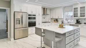 A modern kitchen with white cabinetry, a stainless steel refrigerator, an island with bar stools, and a large window.