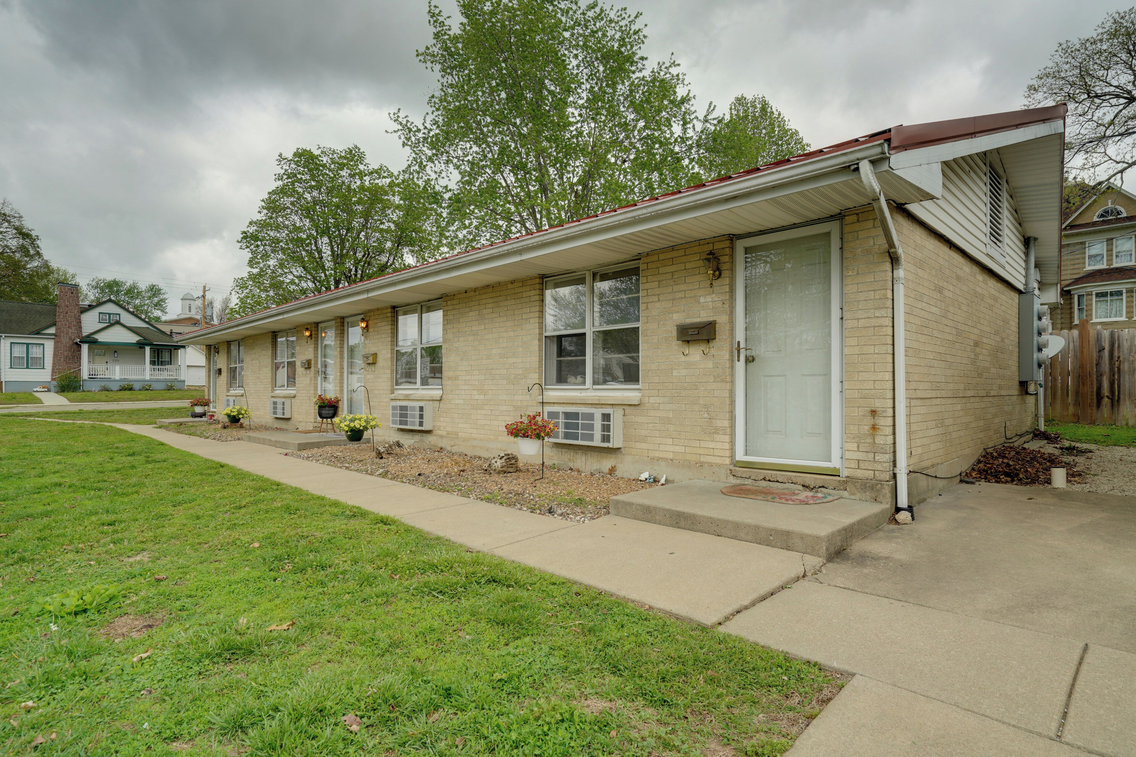 A row of single-story brick houses with attached garages, a grassy lawn, and a cloudy sky.