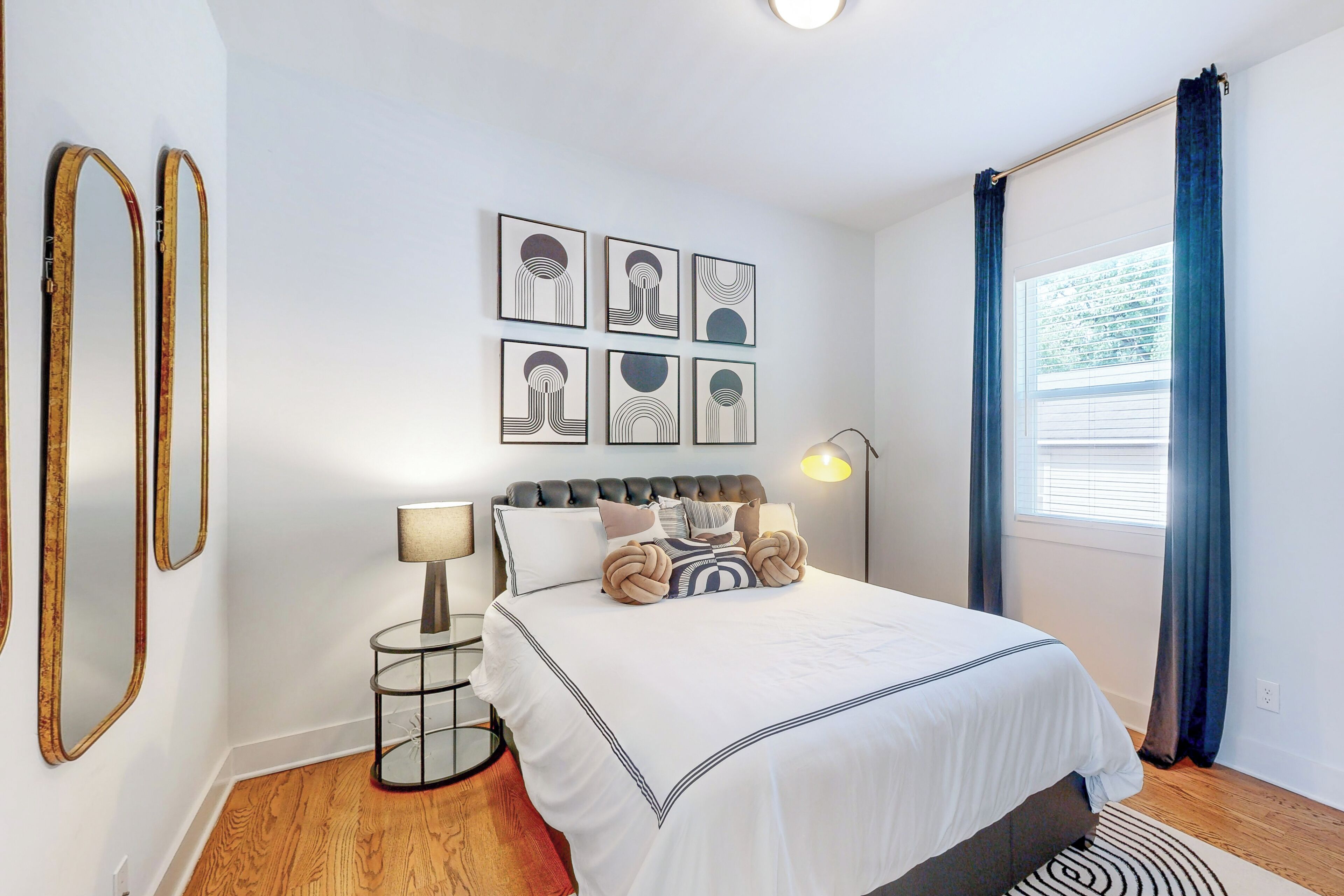 Une chambre à coucher avec un lit, une table de chevet avec une lampe, un miroir fixé au mur et une fenêtre avec des rideaux.