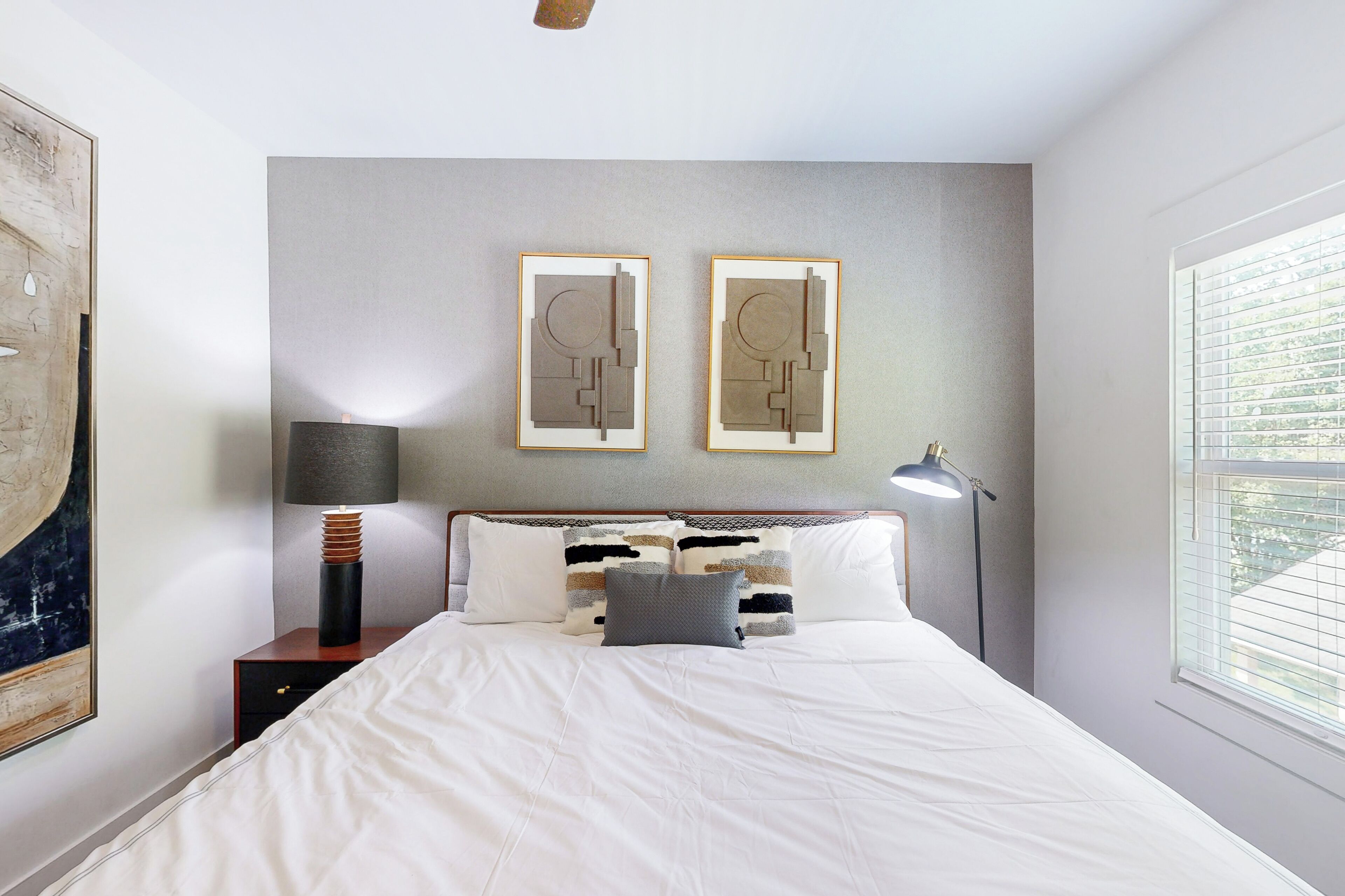 A neatly made bed with white linens, two framed abstract artworks on the wall, a lamp on a bedside table, and a window with blinds.