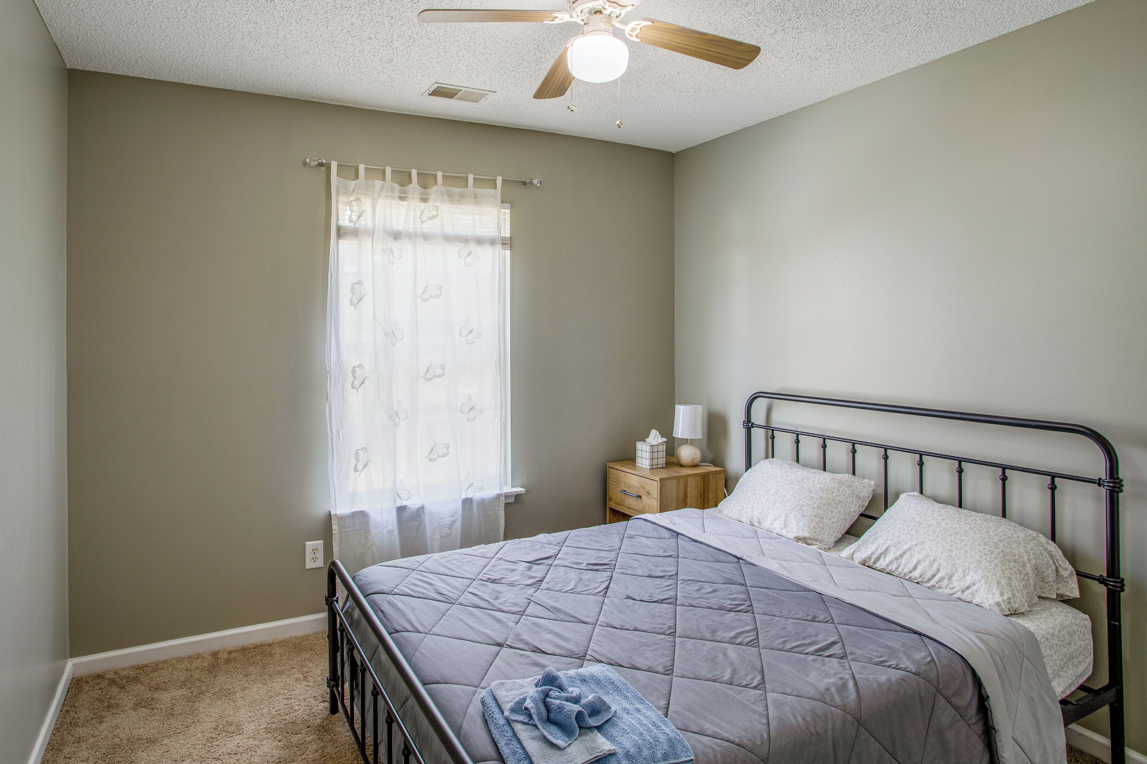 A bedroom with a bed, a nightstand, a ceiling fan, and a window with a curtain.