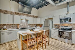 Una cocina con armarios blancos, pared de ladrillo, refrigerador de acero inoxidable, microondas y estufa.