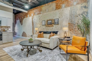 A room with a white sofa, a round table, a yellow armchair, and a brick wall with pictures.