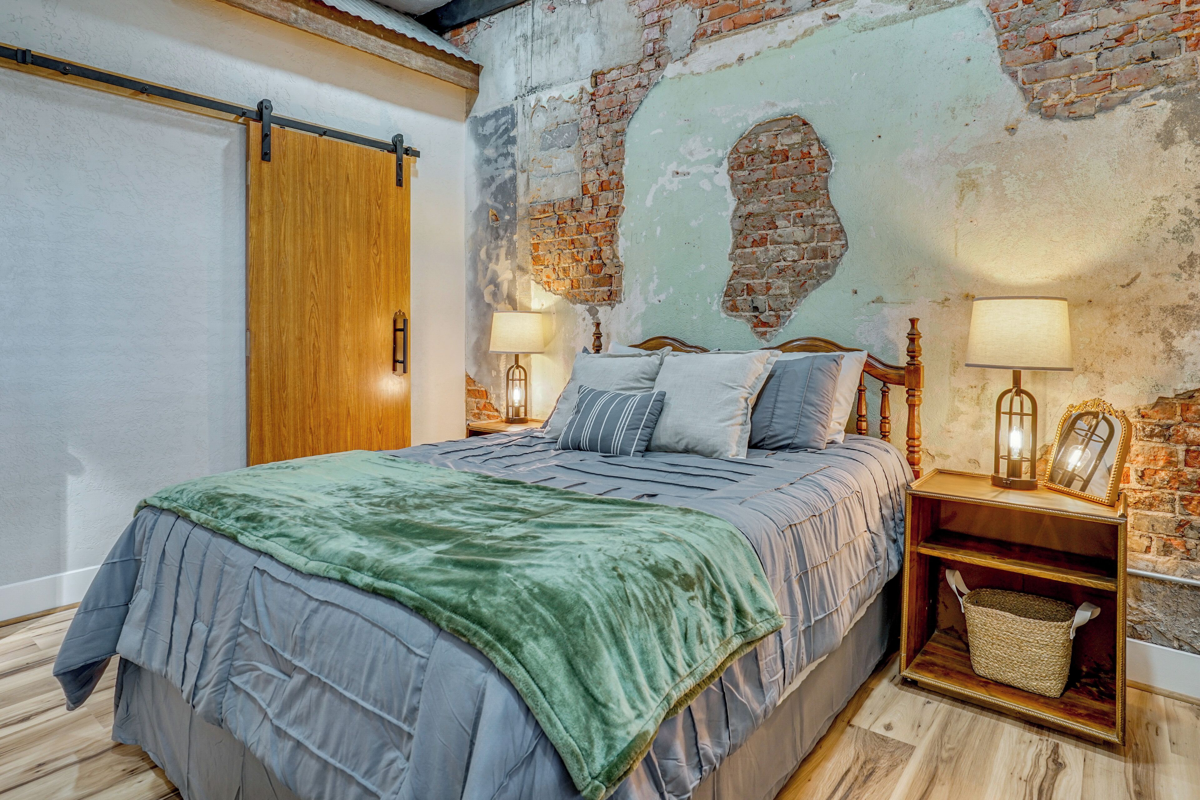 A bedroom with a bed, wooden headboard, two bedside tables with lamps, a wooden nightstand, a basket, and a brick wall with exposed brick.