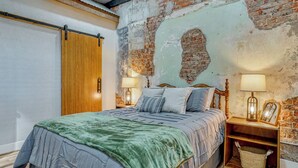 Un dormitorio con cama, cabecera de madera, dos mesitas de noche con lámparas, un pequeño mueble de noche de madera, una cesta y una pared de ladrillo visto.