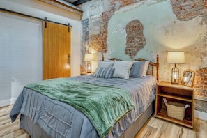 Un dormitorio con cama, cabecera de madera, dos mesitas de noche con lámparas, un pequeño mueble de noche de madera, una cesta y una pared de ladrillo visto.