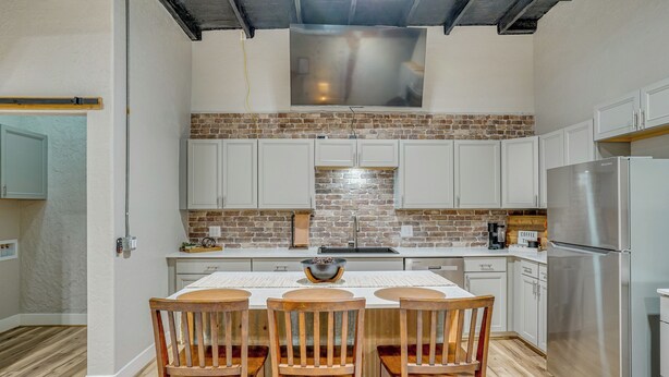 A kitchen with a brick backsplash, white cabinets, a stainless steel refrigerator, and a dining area with wooden chairs and a table.