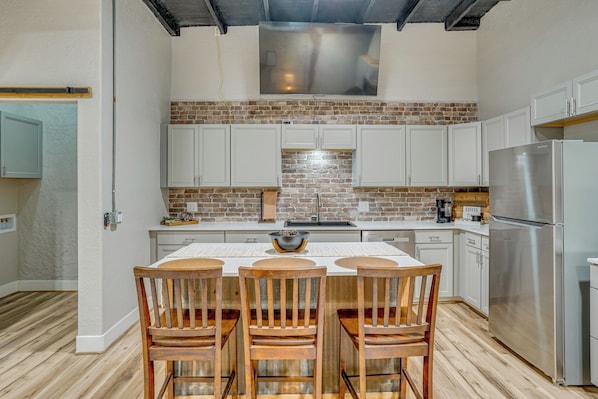 Una cocina con salpicadero de ladrillo, armarios blancos, un refrigerador de acero inoxidable y un área de comedor con sillas de madera y una mesa.