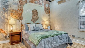 A bedroom with a brick wall, a bed with pillows, a green blanket, wooden nightstands, and two lamps.