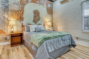A bedroom with a brick wall, a bed with pillows, a green blanket, wooden nightstands, and two lamps.