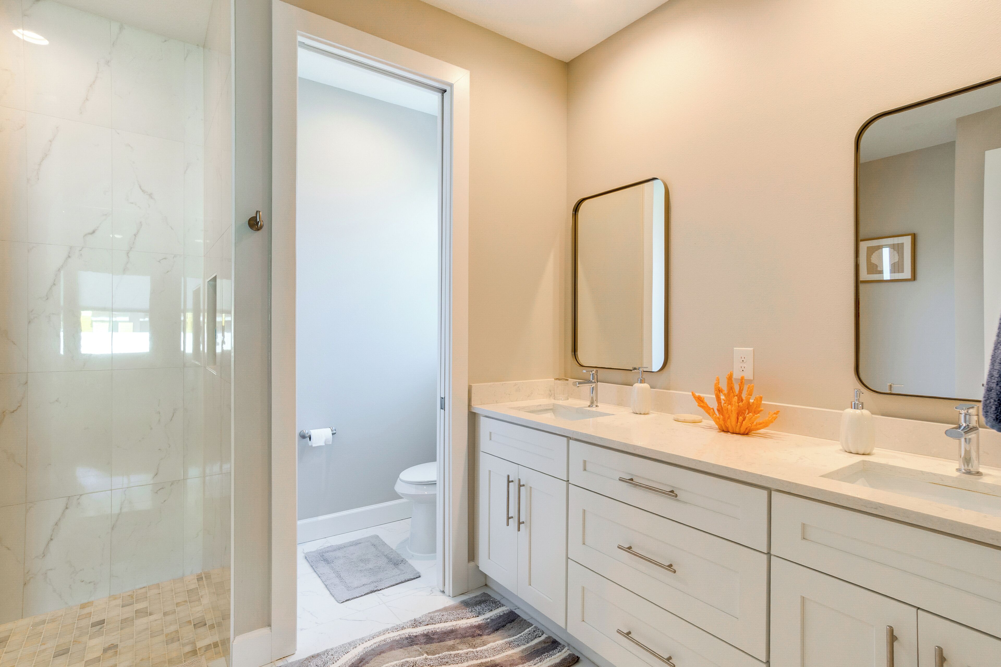 A bathroom with a glass shower enclosure, a toilet, a sink with a mirror, and a rug on the floor.