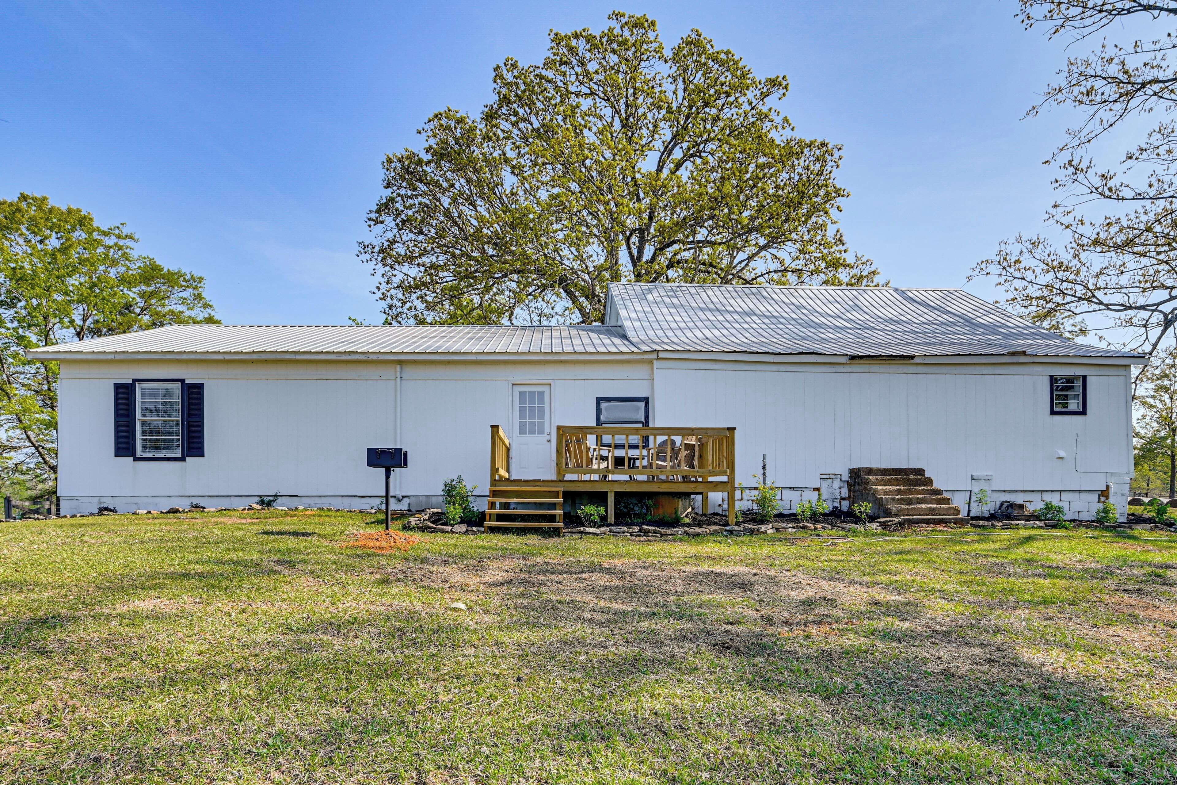 A single-story white house with a metal roof, a wooden deck, and a large tree in the background.